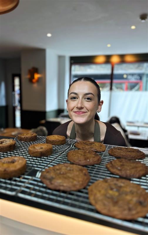 A smiling woman stands behind a wire rack displaying freshly baked cookies in a cozy café setting.