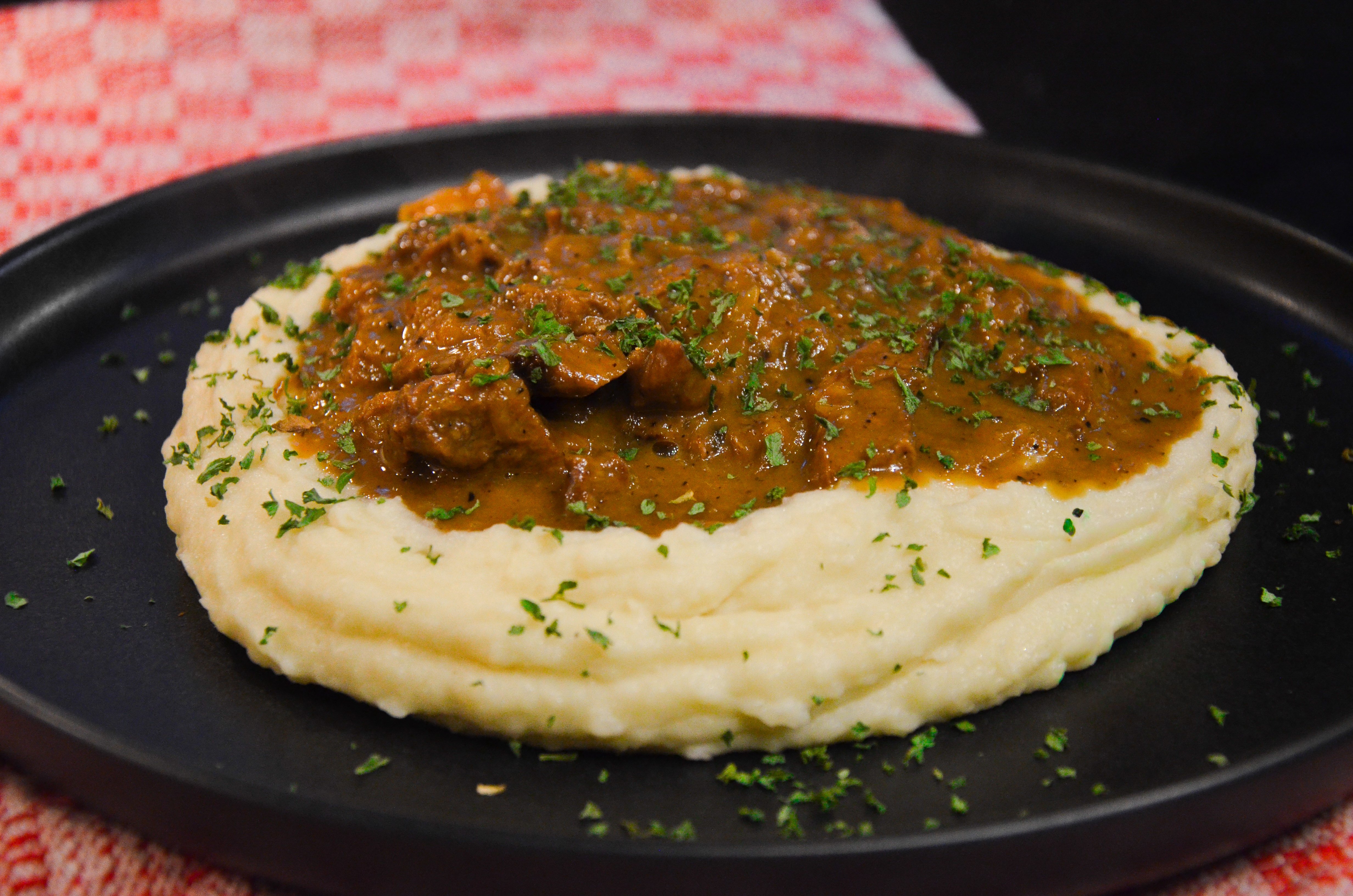 A black plate with creamy mashed potatoes topped with rich beef stroganoff, garnished with fresh parsley. The dish is served on a red and white checkered cloth.